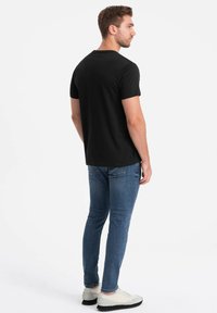 Sort t-shirt med korte ærmer, tætsiddende design. Sammen med blå jeans og lyse sneakers. Baggrunden er ensfarvet hvid.