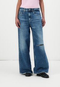 Jeans din denim albastru, largi, cu o nuanță deschisă, având un detaliu rupt la genunchi, bucle pentru curea și buzunare laterale. Purtați cu pantofi negri.