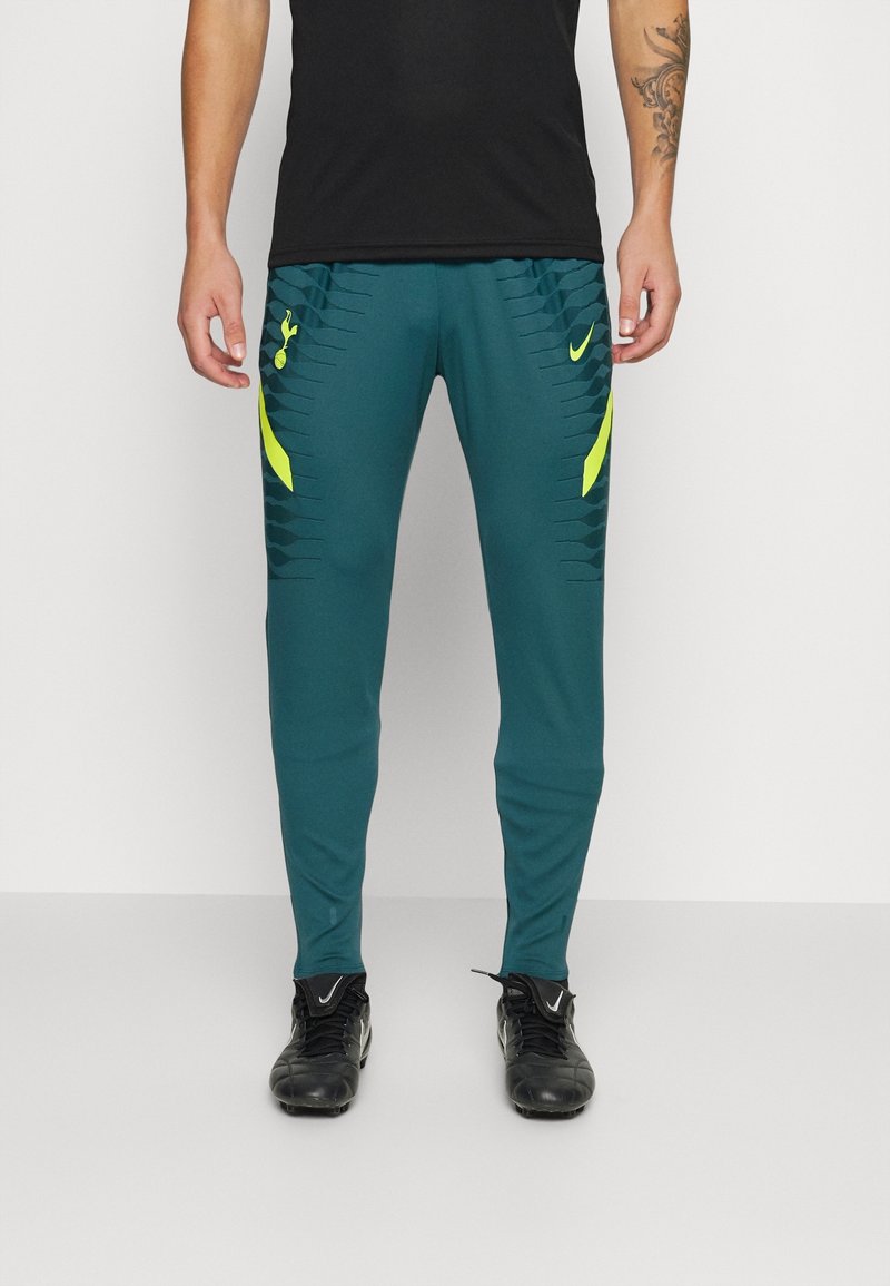 Homme portant un pantalon de soccer Nike Tottenham Hotspur vert sarcelle avec des accents jaunes et des crampons noirs, debout devant un fond blanc.