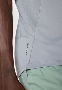 Camiseta deportiva DRI-FIT de color gris claro con tejido texturizado y patrón perforado, combinada con pantalones cortos de color verde menta. Primer plano de la vista lateral.