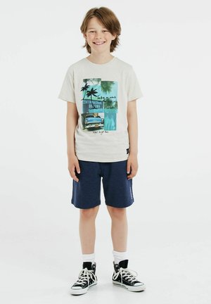 T-shirt chiara con stampa grafica che presenta palme e auto d'epoca, abbinata a pantaloni corti blu navy e sneakers alte nere.