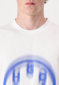 White cotton t-shirt featuring a blue graphic design with dotted pattern, round collar, and short sleeves. Close-up of chest area.