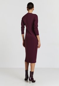 Frau steht mit dem Rücken zur Kamera und trägt ein eng anliegendes, langärmliges, knielanges Kleid in Pflaumenfarbe, passende Knöchelstiefel und goldene Creolen.