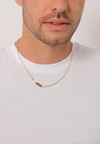 Guld kæde halskæde med aflange led og et rektangulært vedhæng indgraveret med "Kluziol," båret over en simpel hvid t-shirt.