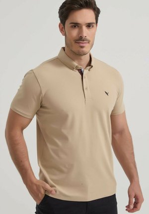 Homme aux cheveux courts et foncés portant un polo beige et un pantalon noir, debout avec une main dans la poche sur un fond uni.