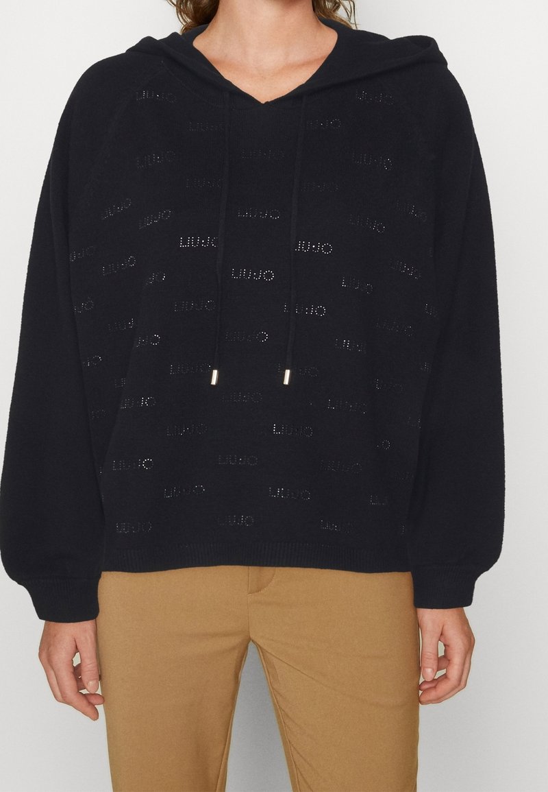 Femme portant un pull à capuche noir avec des motifs discrets du logo « Liu Jo », associée à un pantalon marron clair, sur un fond uni.