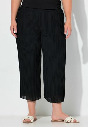 Unterkörper einer Person, die schwarze plissierte, weit geschnittene, gekürzte Hosen und weiße flache Slide-Sandalen mit geflochtenen Riemen trägt.
