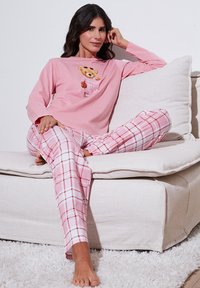 Roza pajkama z dolgimi rokavi z motivom medvedka, kombinirana s kavbojkami v roza in beli karo vzorcu. Mehka tkanina, sproščen kroj, sedi na kavču.