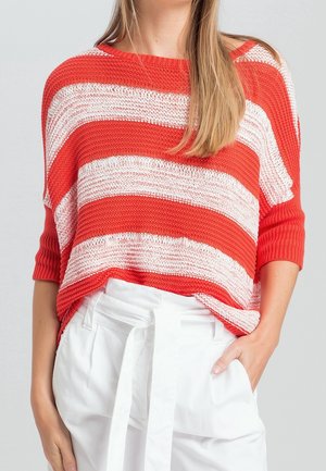 Mujer con un suéter de punto a rayas rojas y blancas con mangas hasta el codo y pantalones blancos de cintura alta con cinturón de tela.