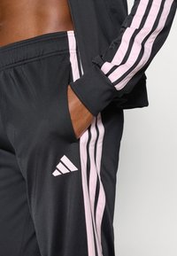 Μαύρη αθλητική φόρμα με ροζ ρίγες ως ακρογωνιαίους λίθους, διαθέτει στενή εφαρμογ ή και ελαστικά μανσέτες. Περιλαμβάνει λογότυπο της Adidas στο πόδι.