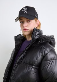 Čierna puff jacket s vysokým golierom a lesklým povrchom, kombinovaná s čiernou baseballovou čiapkou s bielym logom. Pod ňou tmavofialová košeľa.