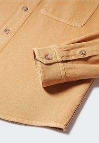 Manchette et ourlet inférieur d'une chemise longue à manches longues couleur beige, boutonnés, avec des coutures visibles et une poche, posée à plat.