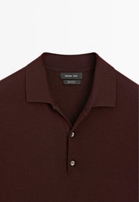 Burgundy bomullspolo med klassisk krage, tvåknapsstängning och strukturerad tyg. Etiketten säger "Massimo Dutti," tillverkad i Kina.