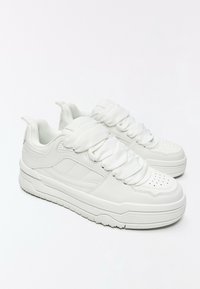 Paire de baskets blanches épaisses avec des lacets épais et texturés, un col matelassé et un détail perforé à l'avant-pied sur un fond blanc uni.