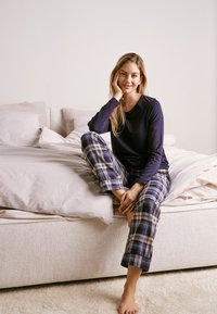 Navy langærmet top parret med ternede pyjamasbukser i blå og beige, siddende på en lys sofa med bløde sengetæpper i baggrunden.
