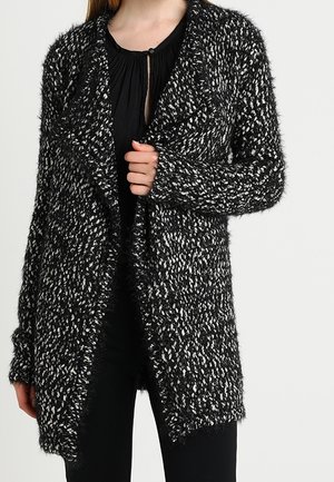 Vrouw die een zwart-witte, gestructureerde cardigan met open voorkant draagt over een zwarte blouse en zwarte broek, staand tegen een effen achtergrond.