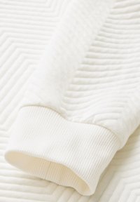 Tessuto bianco a coste con un motivo diagonale testurizzato. Il polsino visibile presenta una finitura morbida ed elastica. Colore uniforme e liscio in tutto il tessuto.