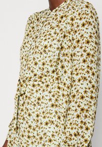 Abito a maniche lunghe di colore verde chiaro con stampa floreale in marrone e giallo. Presenta un colletto con bottoni e una vita con cintura per definire la silhouette.