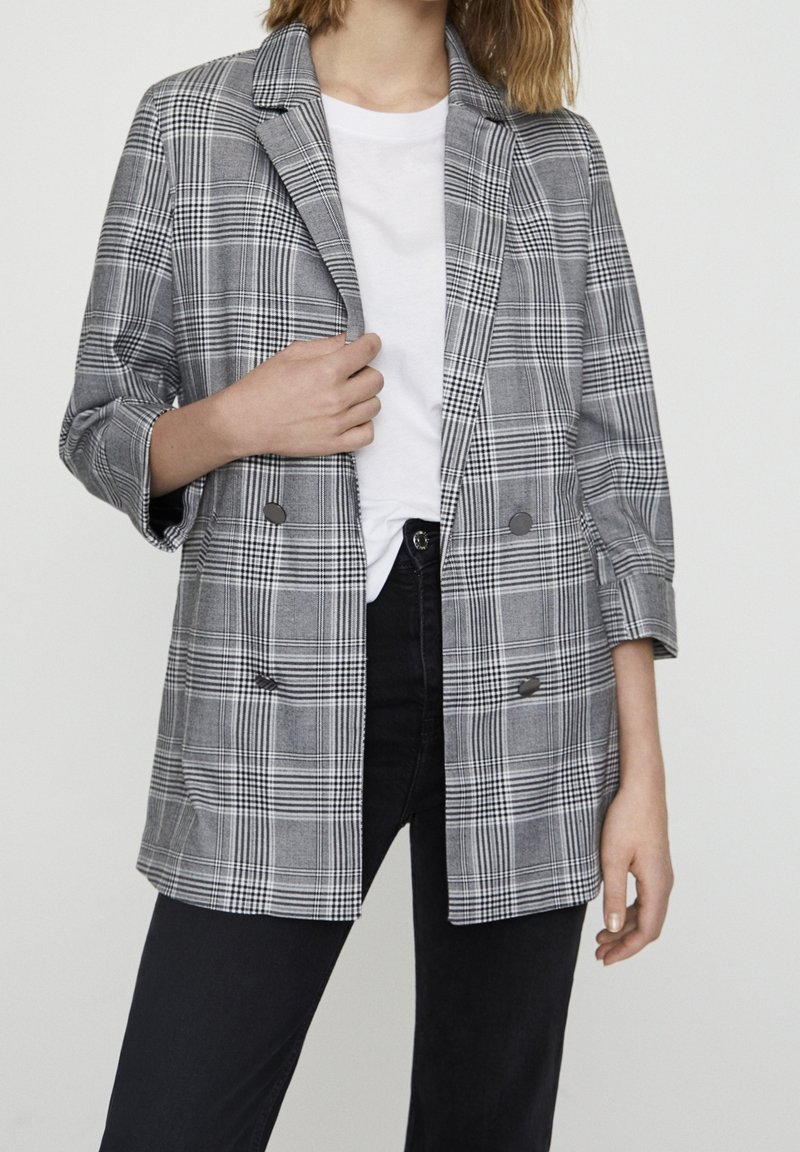 Femme portant un blazer croisé à carreaux gris sur un t-shirt blanc et un jean noir, tenant le revers du blazer d'une main.