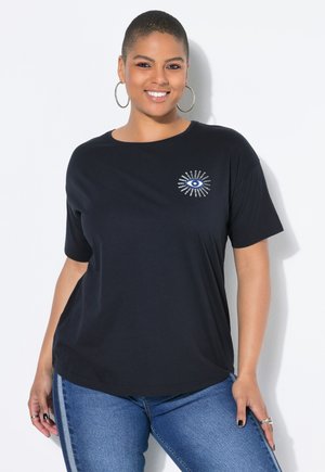 Femme aux cheveux courts portant des boucles d'oreilles créoles, un t-shirt noir avec un graphique d'œil, et un jean bleu, souriant sur fond blanc.