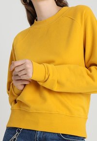 Sudadera amarilla con cuello redondo, puños acanalados y dobladillo. Mangas raglán y textura suave. Vista frontal mostrando las manos cerca de la cintura.