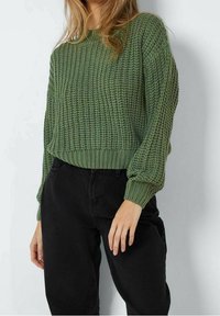 Grüner gestrickter Pullover mit strukturierter Musterung, rundem Ausschnitt und gerippten Bündchen. Kombiniert mit schwarzen Hosen in entspannter Passform.