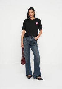 Μαύρο T-shirt με ροζ καρδιά στο σχέδιο, άνετη εφαρμογή, σε συνδυασμό με ψηλόμεσο μπλε τζιν καμπάνα και μαύρα μυτερά φλατ παπούτσια.