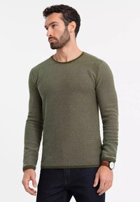 Grünes Langarm-Shirt aus strukturiertem Stoff, mit rundem Ausschnitt und kontrastierender Bordüre. Getragen mit dunklen Jeans und einer Uhr.