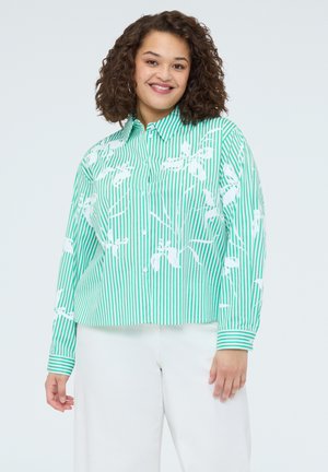 Donna con capelli ricci che sorride, indossa una camicia verde a righe verticali bianche con motivi floreali bianchi e pantaloni bianchi.