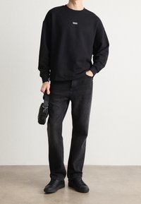 Hombre vistiendo un sudadera negra con el logo "HUGO", jeans negros y zapatillas negras, sosteniendo una pequeña bolsa negra por su asa.