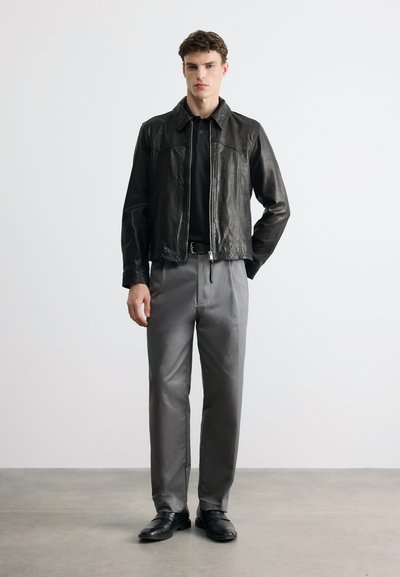Jeune homme debout portant une veste en cuir noire, une chemise noire, un pantalon gris et des chaussures noires sur un fond blanc uni.