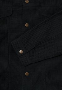 Čierna štruktúrovaná rukávová časť a predná časť bundy s kovovými gombíkmi označenými "BLKVIS" v mriežkovanom vzore.