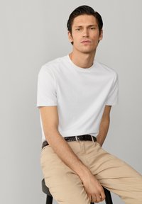 Junger Mann mit dunklen Haaren, der ein weißes kurzärmeliges T-Shirt und beige Hosen trägt, sitzt auf einem schwarzen Hocker vor einem schlichten grauen Hintergrund.