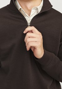 Sudadera con cremallera de color marrón oscuro y cuello alto, textura de tela suave y puños acanalados. Una camisa a rayas blanca es parcialmente visible debajo.