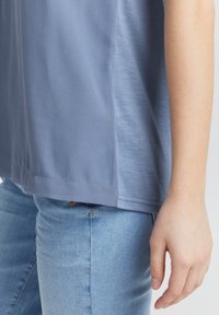 Haut à manches courtes, bleu clair, en tissu lisse ; présente une coupe droite et une texture subtile, porté avec un jean bleu ajusté.