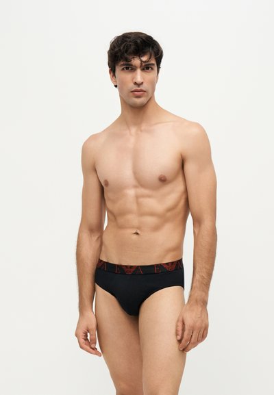Homme torse nu aux cheveux foncés portant un slip noir avec une ceinture rouge à logo, debout devant un fond clair uni.