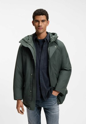 Mand iført en mørkegrøn vinterjakke med hætte over en navy skjorte og sweater med blå jeans, stående mod en ensfarvet lys baggrund.