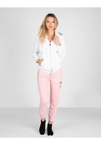 Felpa bianca con zip e cappuccio regolabile, abbinata a pantaloni della tuta rosa con stampa grafica e sneakers nere senza lacci.