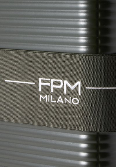 Sangle de bagage texturée grise avec texte blanc "FPM MILANO" enroulée autour d'une surface de valise côtelée foncée.