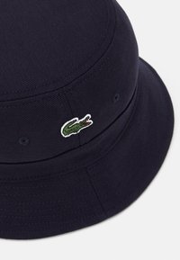 Tamsiai mėlynas kibirinis skrybėlė, pagaminta iš tekstūruoto audinio, su mažu žaliu ir raudonu Lacoste krokodilo logotipu šone.