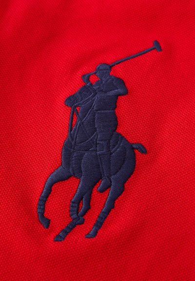 Sarkans audums ar tumši zilu izšūtu polo spēlētāja logo, kas attēlo jātnieku uz zirga, uzrādot paaugstinātu tekstūru un detalizētu šūšanu.