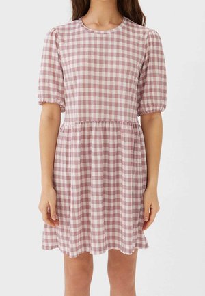 Vrouw die een korte, roze-wit geruite jurk draagt met pofmouwen en een geplooide taille, staand tegen een effen achtergrond.