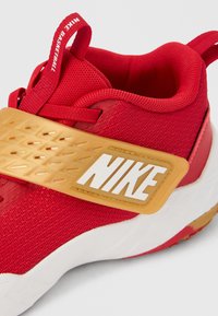 Κόκκινο υφασμάτινο αθλητικό παπούτσι με διαμορφωμένο χρυσό λουράκι με λευκή επιγραφή «NIKE», επενδυμένο κολάρο και υφασμένη ελαστική σόλα.