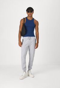 Námořnický top bez rukávů, šedé tepláky s kapsami na zip, bílé tenisky a černá křížová taška. Model stojí proti neutrálnímu pozadí.