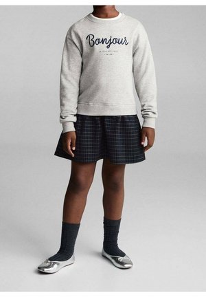 Enfant portant un sweat-shirt gris clair avec le texte "Bonjour", une jupe à carreaux foncés, des chaussettes foncées et des ballerines argentées, debout devant un fond gris.