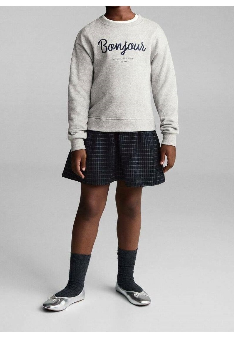 Enfant portant un sweat-shirt gris clair avec le texte "Bonjour", une jupe à carreaux foncés, des chaussettes foncées et des ballerines argentées, debout devant un fond gris.