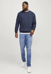 Námořnicky modrá mikina s kulatým výstřihem, žebrovanými manžetami a lemem, vrstvená přes bílou košili, ve spojení s lehkými džínami a bílými teniskami.