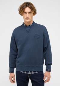 Sweat-shirt bleu marine avec un col rond, des poignets côtelés et un logo circulaire texturé sur la poitrine. Superposé à une chemise à carreaux.