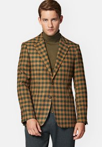 Brązowa i zielona marynarka w kratę z klapami o prostym kroju, z jednym guzikiem i teksturowanym materiałem. Noszona na ciemnym golfie.