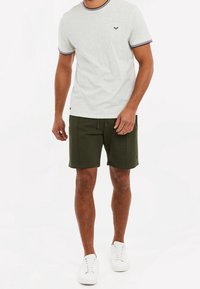 Homme portant un t-shirt à manches courtes gris clair avec des bordures noires, un short vert foncé avec cordons de serrage, et des baskets blanches sur un fond blanc.